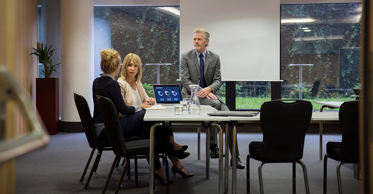 Two women seated at a table with paperwork and a laptop in front of them, a man standing to their right. They're collaborating on a project in a board room.