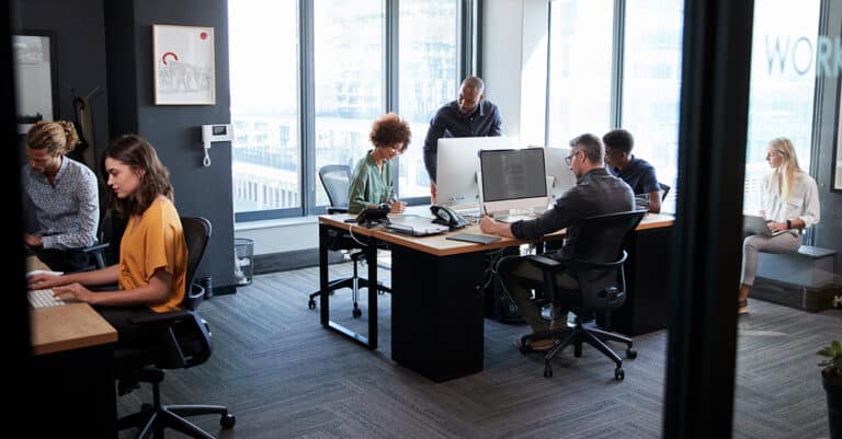 A modern open-plan office with large floor-to-ceiling windows and a group of diverse young professionals working collaboratively. Several people are seated at desks using desktop computers, while others are engaged in conversation or using laptops. The space is well-lit with natural light and features minimalist décor, ergonomic chairs, and carpeted flooring.